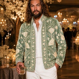 May include: A sage green velvet jacket featuring intricate cream-colored floral embroidery along the front panels and collar, worn over a crisp white button-down shirt and matching white trousers, creating a coordinated ensemble with long sleeves.