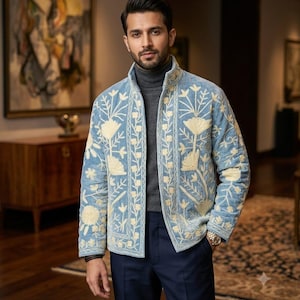 May include: A light blue velvet jacket featuring cream-colored floral embroidery along the front panels and sleeves, worn over a dark gray turtleneck sweater and paired with dark navy blue trousers, creating a structured and layered appearance.
