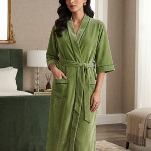 May include: A sage green velvet robe featuring a shawl collar, three-quarter length sleeves, and a matching tie belt at the waist. The garment includes two front patch pockets, each accented with dark green piping along the edges and collar.