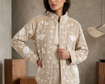 May include: A beige velvet jacket featuring a high collar and long sleeves, adorned with intricate white floral embroidery patterns along the front panels, sleeves, and hemline. The jacket is worn over a plain cream-colored top and beige trousers.