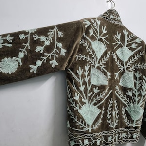 May include: A brown jacket featuring light mint green floral embroidery across the back and sleeves. The garment has a high collar, long sleeves, and a decorative border pattern along the bottom hem, all displayed on a metal hanger.