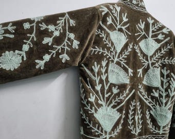 May include: A brown jacket featuring light mint green floral embroidery across the back and sleeves. The garment has a high collar, long sleeves, and a decorative border pattern along the bottom hem, all displayed on a metal hanger.
