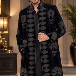 May include: A black velvet long coat featuring a high collar and intricate dark grey floral and circular embroidery along the front placket and sleeves. The garment is worn over a plain black shirt and black trousers, creating a monochromatic look.