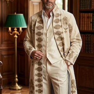 May include: A cream-colored long coat featuring intricate gold floral embroidery along the front panels and cuffs, worn over a matching cream vest, white button-down shirt, and cream trousers. The coat has a high collar and a structured silhouette.