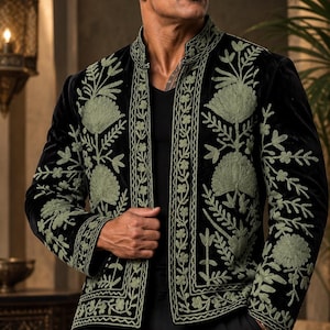 May include: A black velvet jacket featuring a mandarin collar and intricate sage green floral embroidery along the front panels, cuffs, and hem. The garment is worn over a black shirt and paired with black trousers, showcasing a structured design.