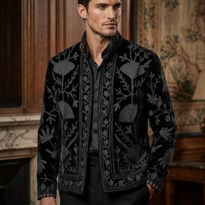 Puede incluir: Una chaqueta de terciopelo negro con cuello alto y mangas largas, adornada con intrincados bordados florales grises en los paneles frontales y los puños. La prenda se lleva sobre una camisa negra abotonada y pantalones negros a juego.