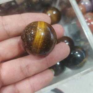 Peut inclure: Une sphère en pierre précieuse œil-de-tigre polie présentant une surface lisse et arrondie avec des bandes chatoyantes brun doré caractéristiques et des inclusions brun foncé, offrant un éclat réfléchissant et un aspect fibreux.