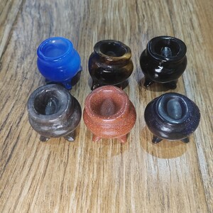 May include: Six small, three-legged cauldron-shaped holders carved from various polished stones. The collection includes one bright blue, one brown tiger eye, one solid black, one grey marbled, one reddish-brown glittery, and one dark blue goldstone.
