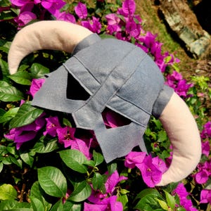 Puede incluir: Un casco de tela gris con dos grandes cuernos curvos de color beige claro que sobresalen de los lados. El casco tiene un diseño estructurado con una banda vertical central y dos aberturas triangulares para los ojos, hecho de fieltro.