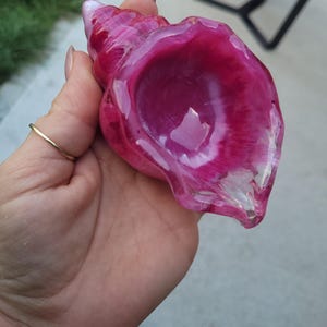 Op de afbeelding: Een decoratief schelpbeeldje gemaakt van doorschijnende hars in levendige tinten magenta en donkerroze. Het object heeft een gladde, glanzende afwerking met wervelende kleurpatronen die de natuurlijke spiraalstructuur van een schelp nabootsen.