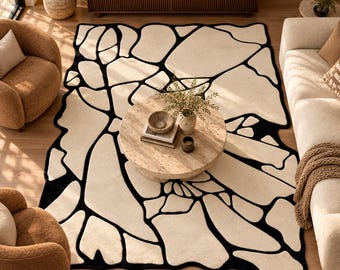 May include: A rectangular area rug featuring an abstract pattern of large, irregular off-white shapes separated by thick black lines. The design resembles a fragmented stone or puzzle-like motif, covering the floor space beneath a round coffee table.