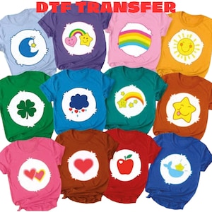 May include: A collection of twelve short-sleeved t-shirts in various solid colors, each featuring a white circular patch on the chest with a unique graphic icon, including hearts, stars, a rainbow, a sun, a clover, a cloud, and an apple.