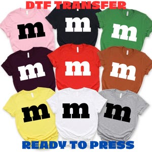 May include: A collection of nine short-sleeved t-shirts in various colors including pink, black, green, dark purple, red, brown, yellow, white, and gray. Each shirt features a large lowercase letter m printed in either black or white on the front.