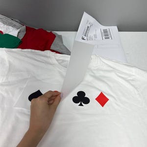 Może przedstawiać: Biały t-shirt z nadrukiem graficznym symboli kart do gry: czarnego pika, czarnego trefla i czerwonego karo. Biały arkusz papieru transferowego jest odklejany od powierzchni koszulki, odsłaniając nadrukowany wz&oacute;r znajdujący się pod spodem.