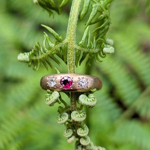 May include: A gold band ring featuring a central round-cut deep pink gemstone flanked by two smaller round-cut clear white gemstones. The stones are set into the metal band, which has a smooth, matte finish and a rounded profile.