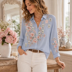 May include: A light blue button-down shirt featuring a floral print of pink cherry blossoms and scattered mahjong tiles. The shirt has long sleeves, a classic collar, and is tucked into white trousers with a brown leather belt.