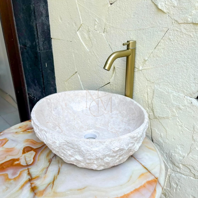 Puede incluir: Un lavabo de piedra beige de forma redonda con una textura exterior rugosa y natural y un interior liso. El lavabo cuenta con un orificio de drenaje central y se combina con un grifo cil&iacute;ndrico alto de metal dorado cepillado.