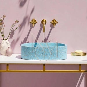 Puede incluir: Un lavabo ovalado de terrazo azul claro con motas multicolores se asienta sobre una encimera blanca. Sobre el lavabo hay un grifo dorado montado en la pared con dos mandos en cruz. Un jarrón blanco con flores y una jabonera están presentes.