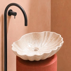 May include: A beige, shell-shaped stone vessel sink with a textured, scalloped rim and a central drain hole. The sink is paired with a tall, matte black curved faucet featuring a single side handle, positioned against a solid peach-colored wall.