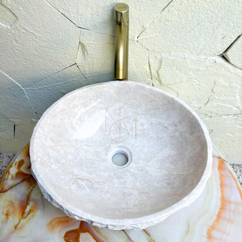 Puede incluir: Un lavabo de piedra redondo en color beige claro con un exterior texturizado natural y un interior liso. El lavabo cuenta con un orificio de drenaje central y se combina con un grifo cil&iacute;ndrico alto de metal dorado cepillado detr&aacute;s.
