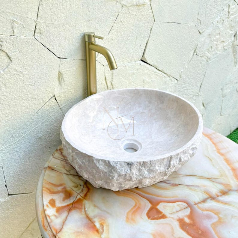 Puede incluir: Un lavabo de piedra beige de forma redonda con un exterior de textura rugosa y natural y un interior liso. El lavabo cuenta con un orificio de drenaje central y se combina con un grifo alto de oro cepillado sobre una superficie de m&aacute;rmol veteado.