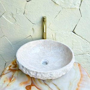Puede incluir: Un lavabo de piedra beige de forma redonda con un exterior de roca natural texturizada y un interior liso. El lavabo cuenta con un orificio de drenaje central y se combina con un grifo met&aacute;lico cil&iacute;ndrico de tono dorado.