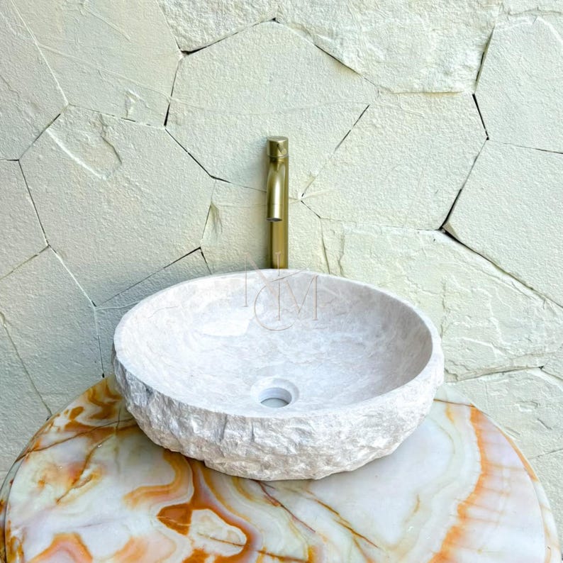 Puede incluir: Un lavabo de piedra redondo en color blanco roto con un exterior rugoso y texturizado y un interior liso. El lavabo cuenta con un orificio de drenaje central y se combina con un grifo met&aacute;lico cil&iacute;ndrico alto en tono oro cepillado.