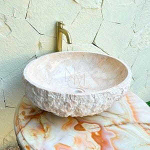 Puede incluir: Un lavabo de piedra beige de forma redonda con una textura exterior rugosa y natural y un interior liso. Un grifo de metal dorado se sitúa detrás del lavabo, el cual está montado sobre una superficie de mármol pulido multicolor.
