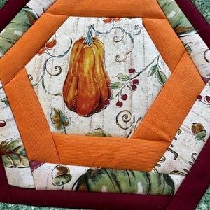 Peut inclure: Un dessous de plat hexagonal en tissu avec un panneau central illustrant une citrouille sur fond crème, entouré de bordures en tissu orange, d'un cadre marron foncé et d'une bordure extérieure en tissu vert à petits motifs floraux blancs.