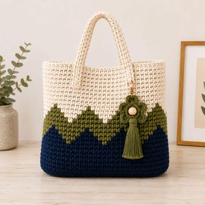 May include: A crochet tote bag featuring a color-blocked design with cream, olive green, and navy blue yarn in a chevron pattern. The bag has two top handles and a decorative olive green flower-shaped charm with a wooden bead and tassel.