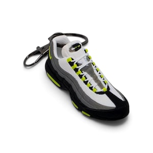May include: A miniature sneaker keychain featuring a layered design in white, grey, and black with vibrant neon yellow accents on the eyelets and sole. The shoe has white laces and a black metal lobster clasp attached to the heel area for hanging.