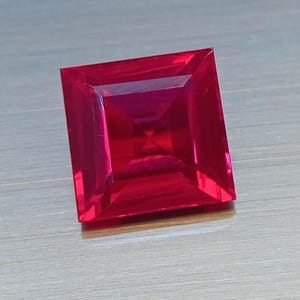 May include: A square-cut, vibrant red gemstone with a faceted surface that reflects light. The stone features a sharp, geometric shape with clear, symmetrical lines and a deep, saturated crimson colour throughout its transparent, polished body.
