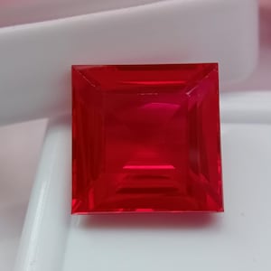 May include: A square-cut, vibrant red gemstone with a faceted surface that reflects light. The stone features sharp, clean edges and a symmetrical geometric shape, displaying a deep, saturated crimson hue throughout its transparent body.