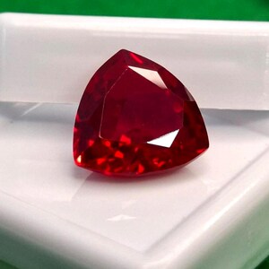 Peut inclure: Une pierre précieuse d'un rouge profond et vibrant, taillée en forme triangulaire avec de multiples facettes qui reflètent la lumière. La pierre présente une teinte cramoisie saturée et brillante ainsi qu'une surface polie et transparente.