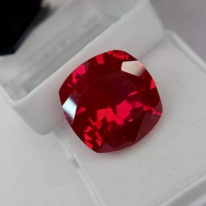 May include: A vibrant, cushion-cut red gemstone with multiple facets that reflect light. The stone is transparent and displays a deep, saturated crimson hue throughout its structure, resting against a white, textured display surface.