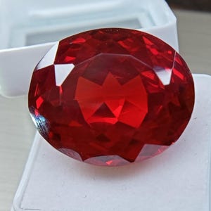Pode incluir: Uma grande pedra preciosa de corte oval com uma cor vermelha vibrante e profunda, apresentando numerosas superfícies facetadas que refletem a luz. A pedra preciosa está posicionada sobre uma caixa de exibição branca aberta com base quadrada.