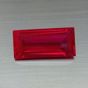 Puede incluir: Una gema de corte rectangular vibrante con un color rojo intenso y saturado. La piedra presenta una superficie facetada que refleja la luz, creando una apariencia clara y transparente con bordes definidos y un acabado pulido y liso.