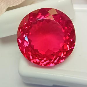May include: A round, faceted gemstone with a vibrant, deep pinkish-red colour. The stone features a precise, symmetrical cut with numerous triangular facets that reflect light, creating a brilliant, sparkling appearance across its surface.