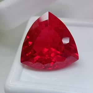 Peut inclure: Une pierre précieuse d'un rouge profond et vibrant, taillée en forme triangulaire avec de multiples facettes qui reflètent la lumière. La pierre présente une teinte cramoisie saturée et une silhouette géométrique aux coins pointus.