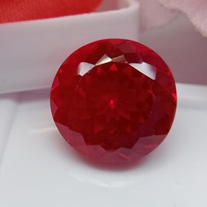Könnte beinhalten: Ein runder, facettierter Edelstein in einer leuchtenden, tiefroten Farbe. Der Stein weist einen komplexen, symmetrischen geometrischen Schliff auf, der zahlreiche dreieckige und rautenförmige Facetten erzeugt, die das Licht reflektieren.