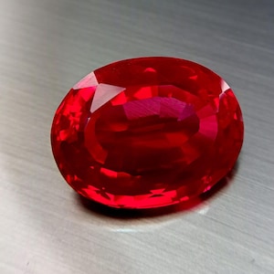 Puede incluir: Una gema de corte ovalado con un color rojo intenso y saturado. La piedra presenta una superficie facetada que refleja la luz, creando un aspecto brillante y centelleante con patrones geométricos definidos en su parte superior.