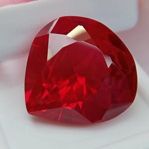 Peut inclure: Une pierre précieuse rouge taillée en forme de cœur avec une finition brillante et transparente. La pierre présente des teintes cramoisies profondes et des facettes géométriques nettes qui reflètent la lumière, créant un aspect poli.