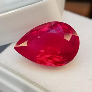 Könnte beinhalten: Ein birnenförmiger, facettierter Edelstein in einer leuchtenden, tiefroten Farbe. Der Stein hat eine polierte Oberfläche mit mehreren geometrischen Facetten, die das Licht reflektieren und für ein brillantes Aussehen sorgen. Er liegt auf einer weißen Unterlage.