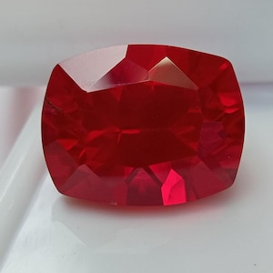 May include: A vibrant, deep red gemstone with a cushion cut, featuring multiple triangular and trapezoidal facets that create a symmetrical, reflective surface. The stone displays a uniform, intense crimson color throughout its transparent body.