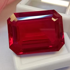 Può includere: Una gemma rosso vivo e profondo con taglio a smeraldo, caratterizzata da sfaccettature nitide e pulite che riflettono la luce sulla sua superficie lucida. La pietra rettangolare è appoggiata sopra una piccola scatola espositiva in plastica bianca.