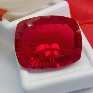 Può includere: Una gemma rosso intenso e vibrante con taglio a cuscino rettangolare e bordi sfaccettati. La pietra è traslucida e presenta una superficie brillante e riflettente, riposta all'interno di una custodia rettangolare bianca e liscia.