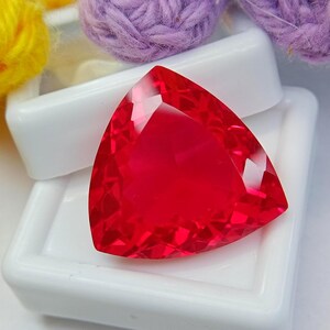May include: A vibrant, transparent red gemstone cut into a triangular shape with multiple facets that reflect light. The gemstone is positioned on a white plastic display box, with blurred yellow and purple yarn balls visible in the background.