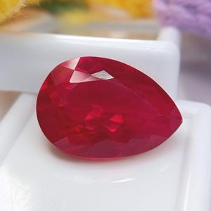 Peut inclure: Une pierre précieuse à facettes en forme de poire d'une couleur rouge profond et vibrante. La pierre présente une surface polie avec de multiples facettes géométriques qui reflètent la lumière, créant un aspect brillant sur fond blanc.