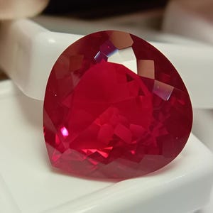 May include: A heart-shaped, faceted gemstone with a deep, vibrant red colour. The stone features numerous geometric cuts across its surface, creating a reflective and brilliant appearance. It is positioned against a smooth, white, glossy surface.