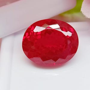 Könnte beinhalten: Ein leuchtender, oval geschliffener Edelstein mit tiefroter Farbe und einer facettierten Oberfläche, die das Licht reflektiert. Der Stein liegt auf einer weißen, glatten, rechteckigen Basis und zeigt seine Klarheit und satte Farbe.